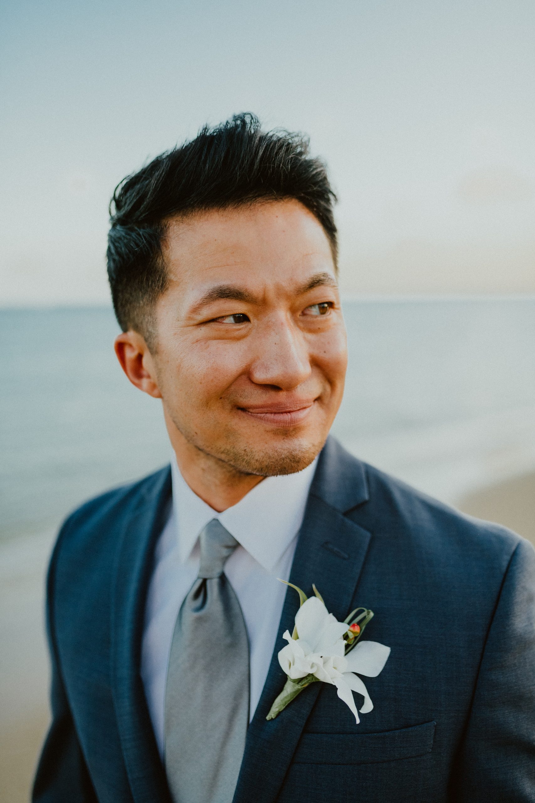 Groom smiles at his bride while wearing his white Hawaiian boutonniere on the lapel of his groom style blue grey suit with silver tie 