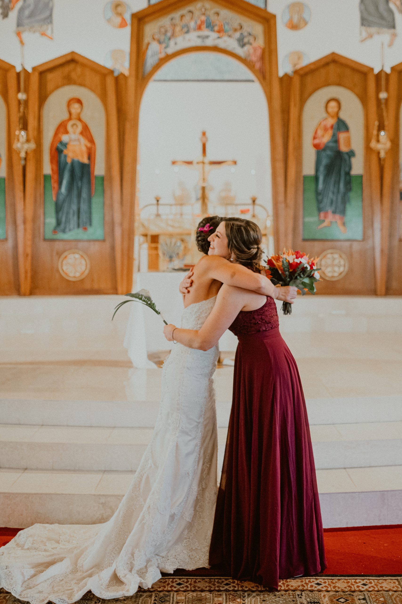 Bride in white lace style dress with a medium-length train hugs her bridesmaid dressed in a maroon colored bridesmaid dress while holding her brightly colored Hawaiian floral bouquet 