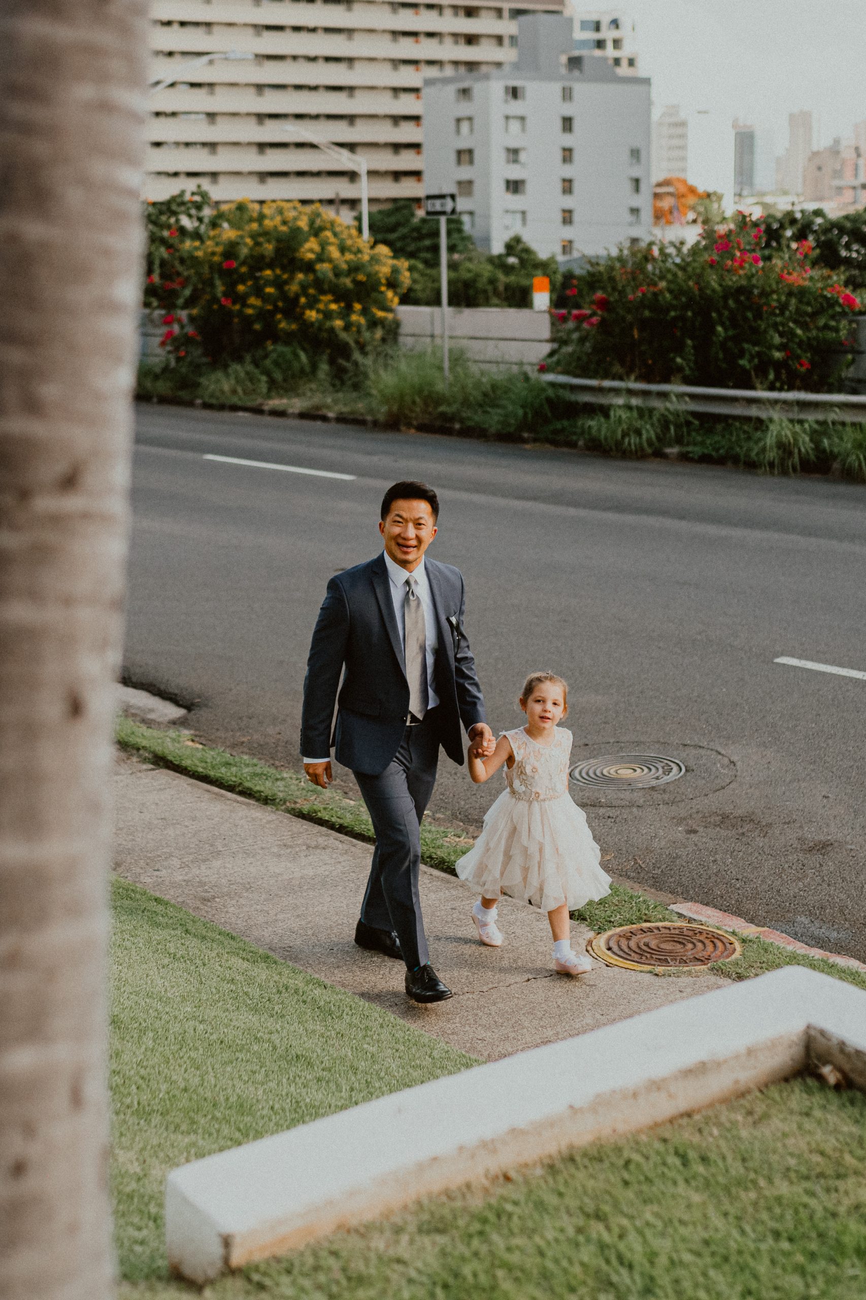 Groom walks down the street of Oahu in front of the church while holding the hand of the flower girl