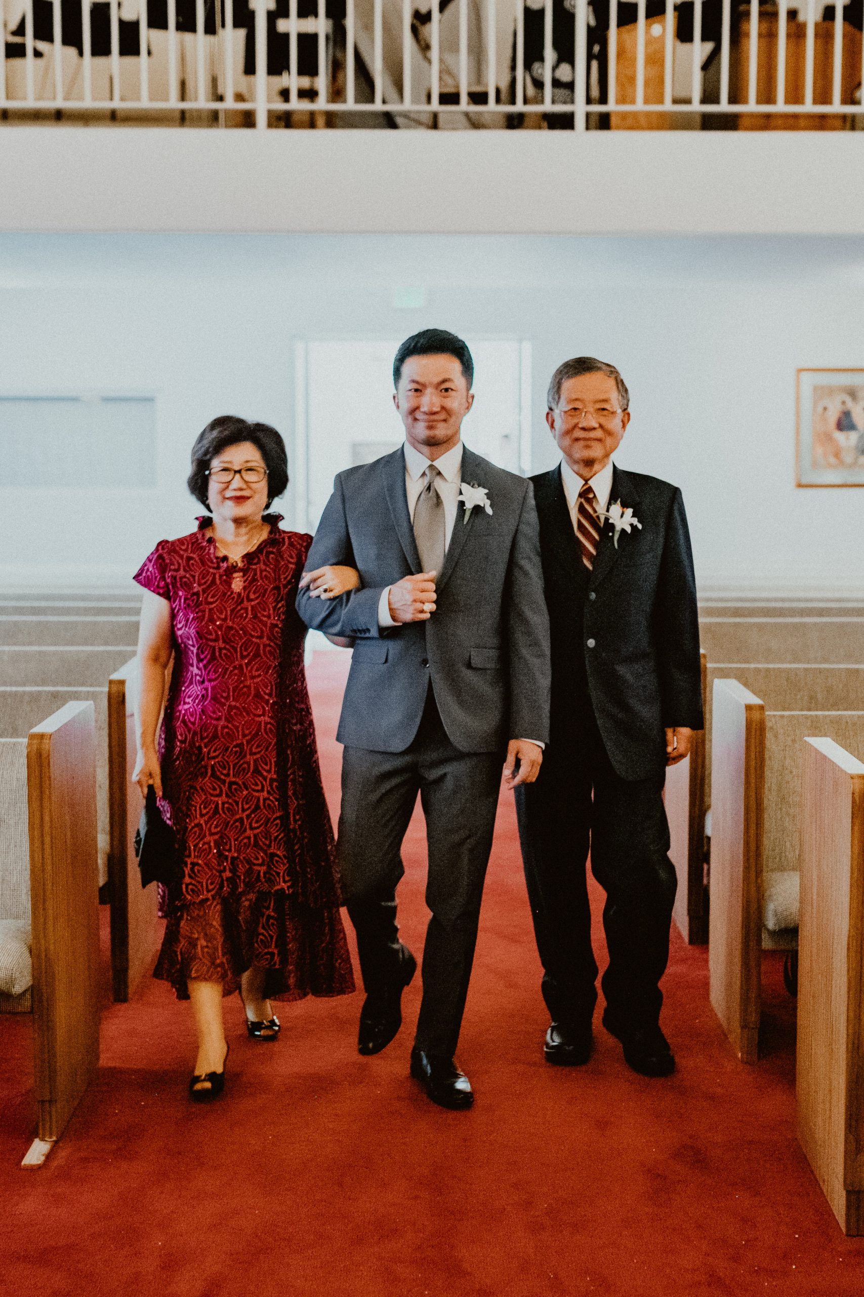 Groom walks down the aisle in a blue-grey suit with his mother wearing a maroon colored dress and his father with a black suit and boutonniere pin 
