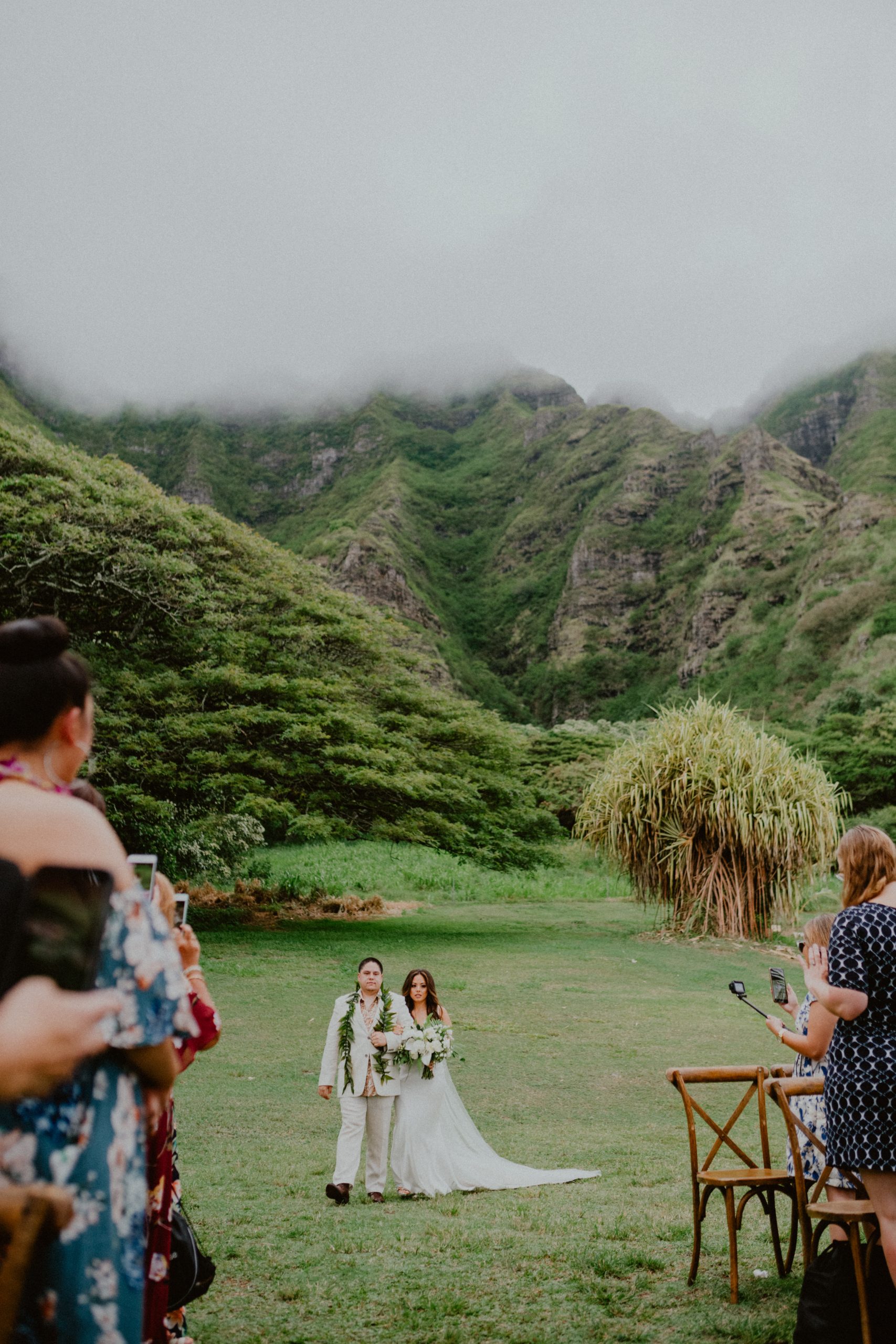 Bride walks down the aisle in front of a gorgeous background of Hawaiian mountains and countryside while wedding guests watch her look at her walking down the aisle | Oahu Wedding Photographer, Oahu Elopement Photographer, Destination Wedding Photographer, Destination Elopement Photographer, Newlywed moments ideas, Newlywed Photography inspiration, Hawaii Elopement ideas | chelseaabril.com