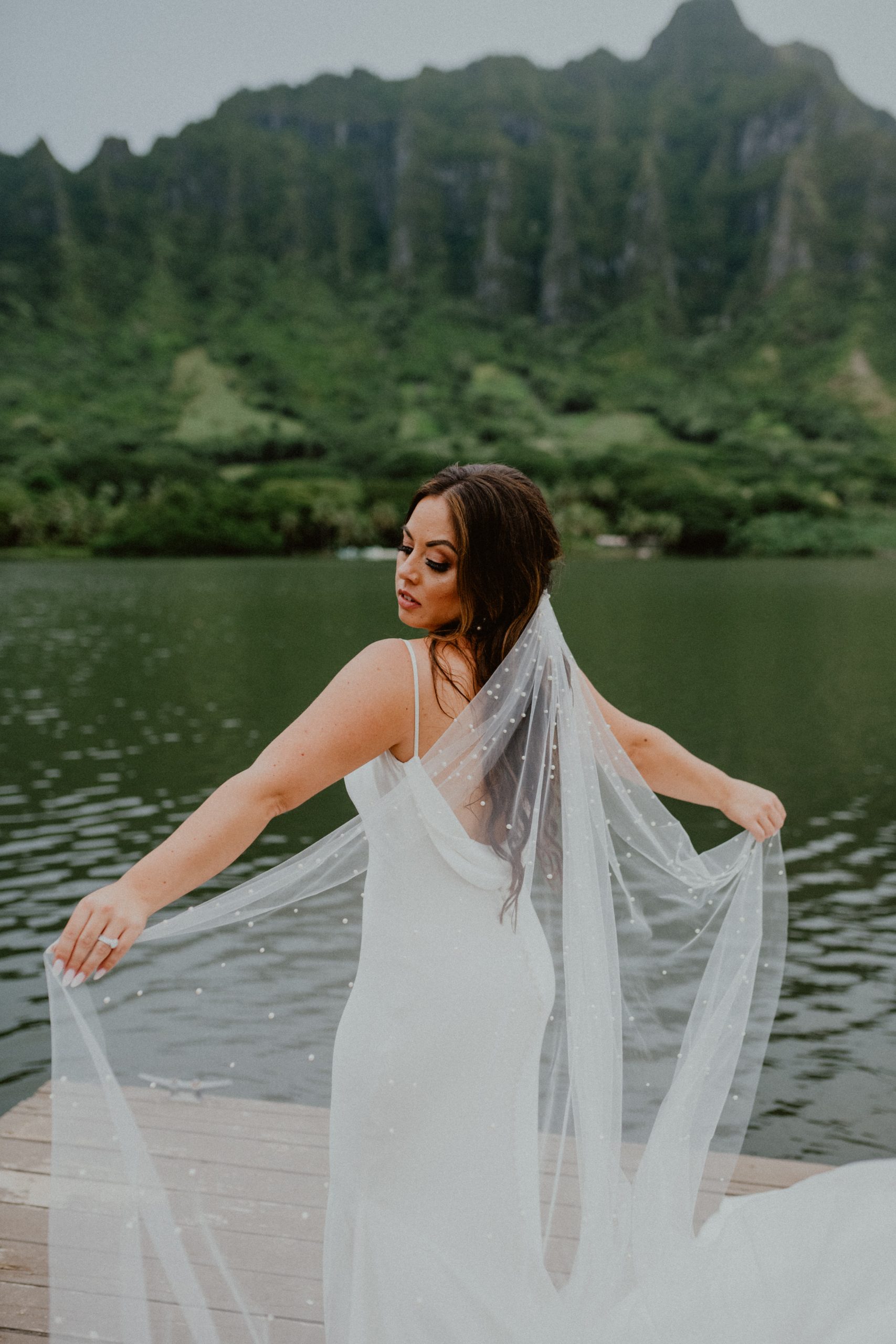 Bride holds out her long veil on the dock of the Kualoa Ranch in Oahu while staring out while wearing her white silk bridal dress | Oahu Wedding Photographer, Oahu Elopement Photographer, Destination Wedding Photographer, Destination Elopement Photographer, Newlywed moments ideas, Newlywed Photography inspiration, Hawaii Elopement ideas | chelseaabril.com