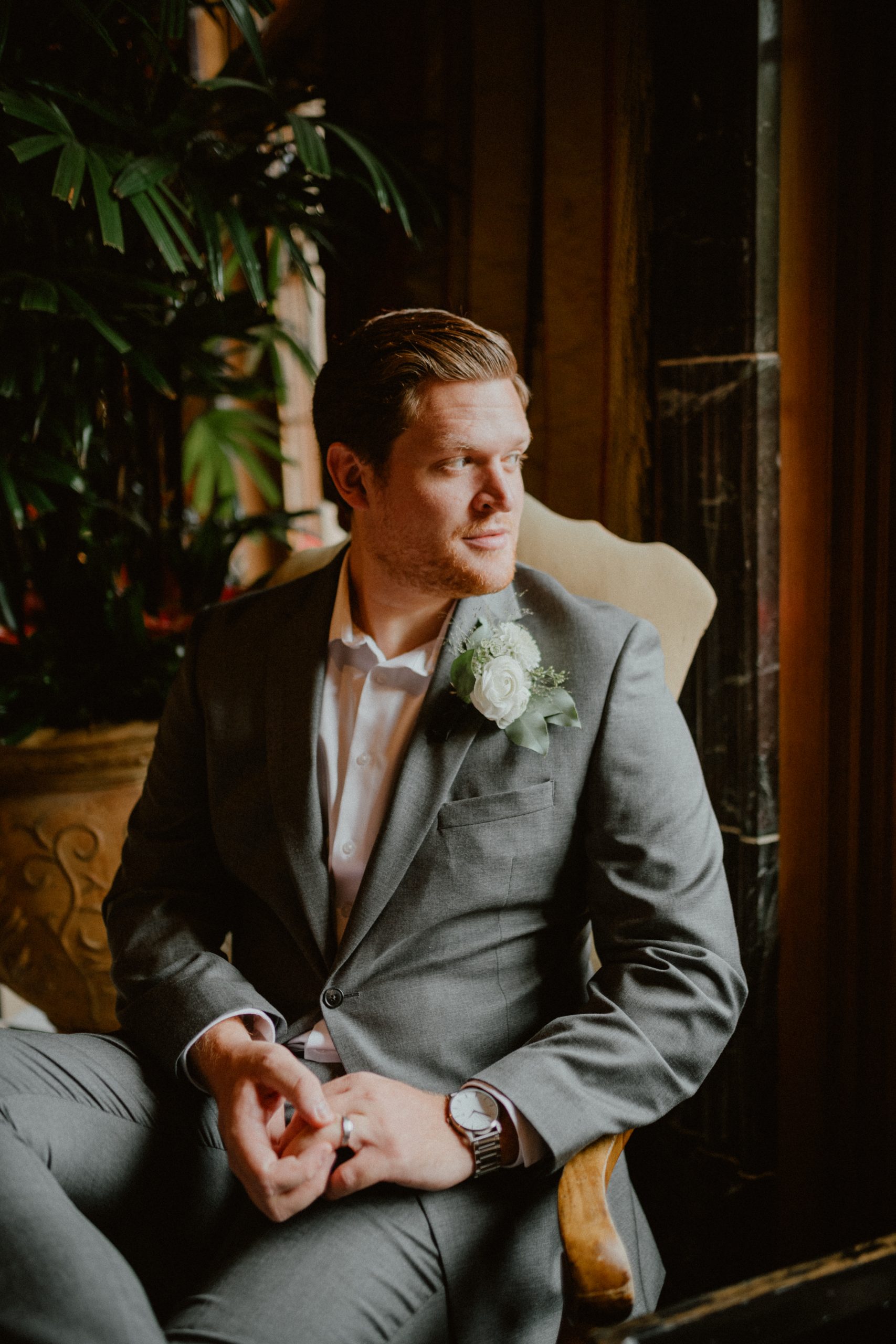 Groom looks out of the hotel window, and wears his grey suit with a white rose boutonniere and a silver watch with his wedding ring | Groom style tips, Groom wedding day inspiration, Groom Tips Wedding, Groom tips for men, Groom tips | chelseaabril.com