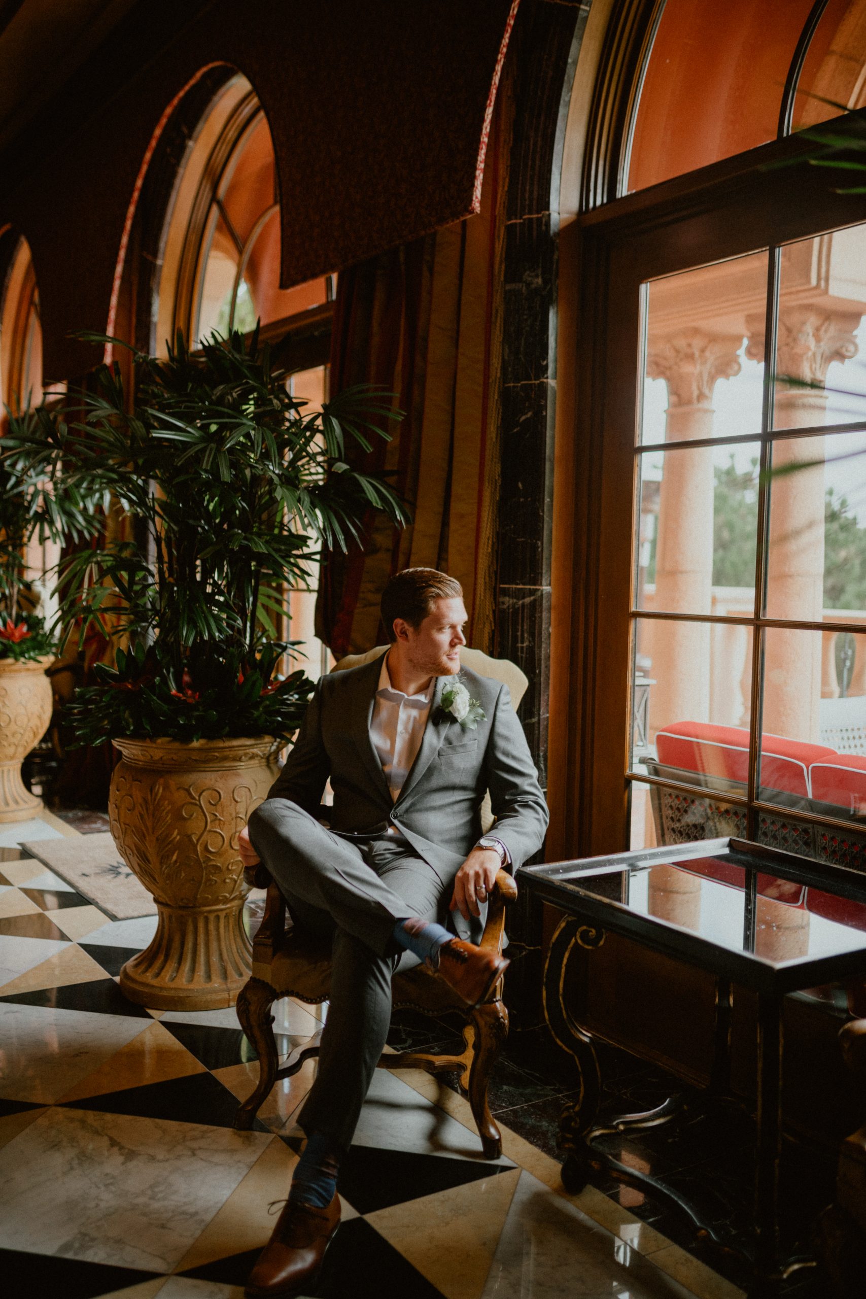 Groom sits in the lobby of the hotel while looking out the window in the hotel lobby, while wearing a grey suit and tan shoes with decorative socks | Groom style tips, Groom wedding day inspiration, Groom Tips Wedding, Groom tips for men, Groom tips | chelseaabril.com
