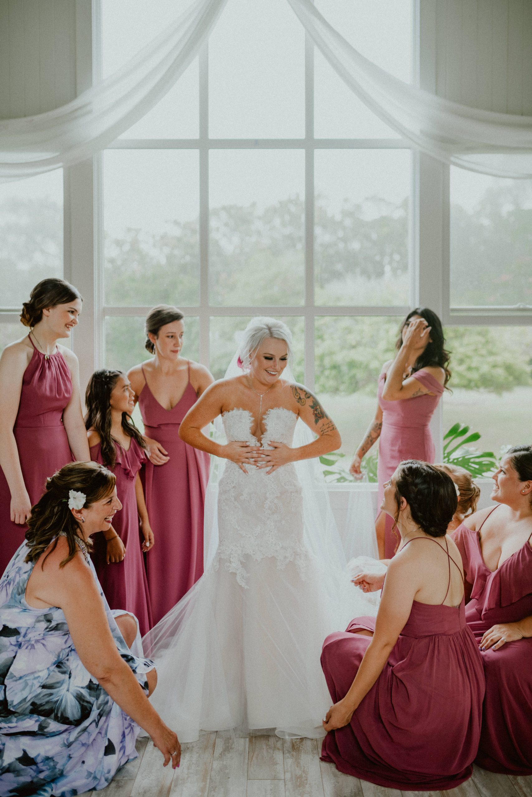 Bride and her bridal party stand in front of large window looking out at Hawaiian Landscape. Bride stands with a mermaid style strapless dress and long tulle dress surrounded by bridesmaids wearing different shades of pink bridesmaid dresses | Oahu Wedding Photographer, Oahu Elopement Photographer, Destination Wedding Photographer, Destination Elopement Photographer, Newlywed moments ideas, Newlywed Photography inspiration, Hawaii Elopement ideas | chelseaabril.com