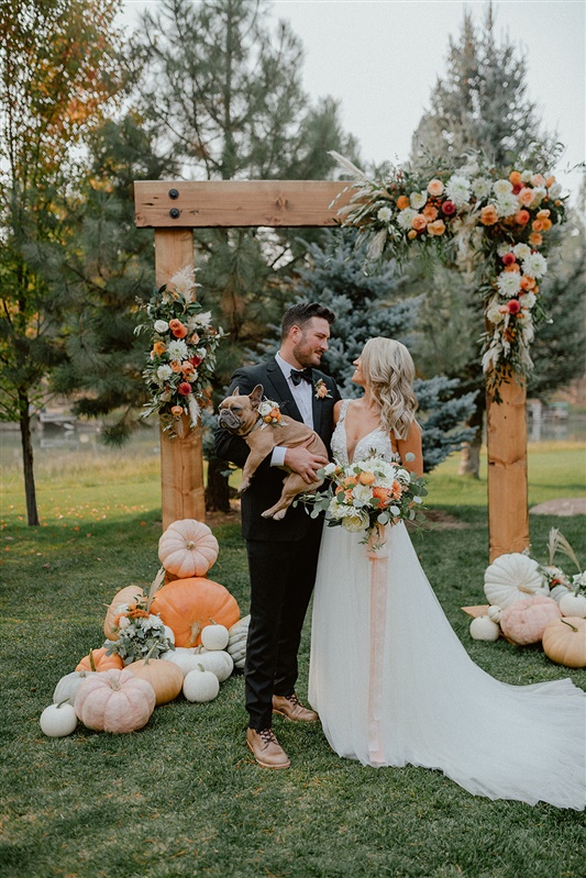 Bride and groom smile at each other while holding their puppy with bow tie in front of Fall inspired wedding altar holding orange and Fall color wedding floral inspiration | Sun River Elopement, Destination Elopement Photographer, Smith Rock Elopement Inspiration, Oregon Elopement ideas, PNW Fall Wedding Inspiration, Fall Outdoor Wedding Ideas,Fall Wedding Inspiration, PNW Outdoor Wedding Ideas | chelseaabril.com