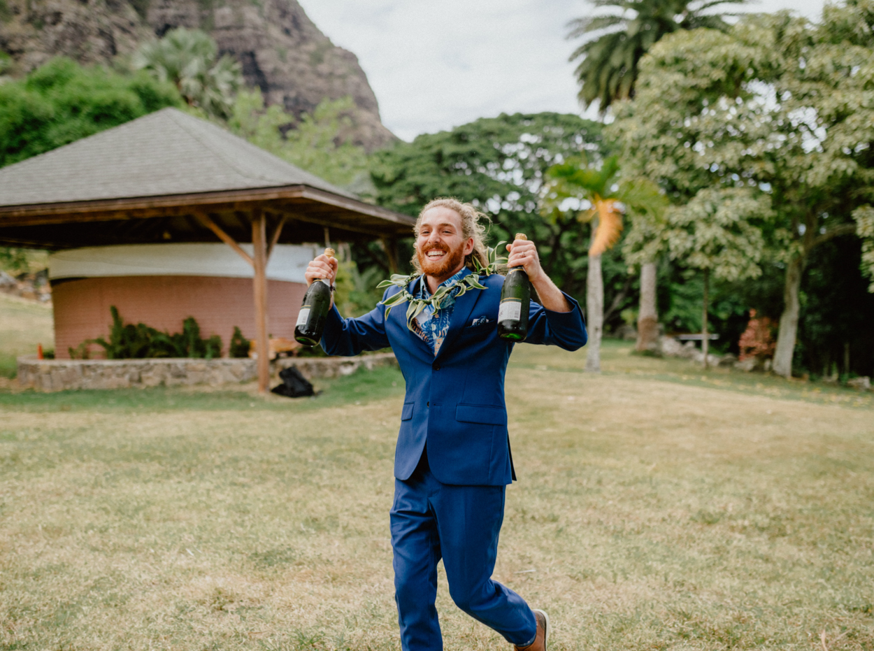 Happy groomsman with champagne in Paliku Gardens Venue at Kualoa Ranch wedding with Chinaman's hat background